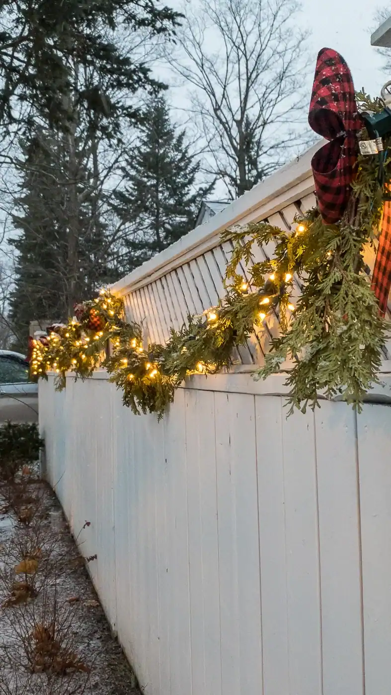 Lit Christmas garland swagged along a white fence with buffalo plaid bows