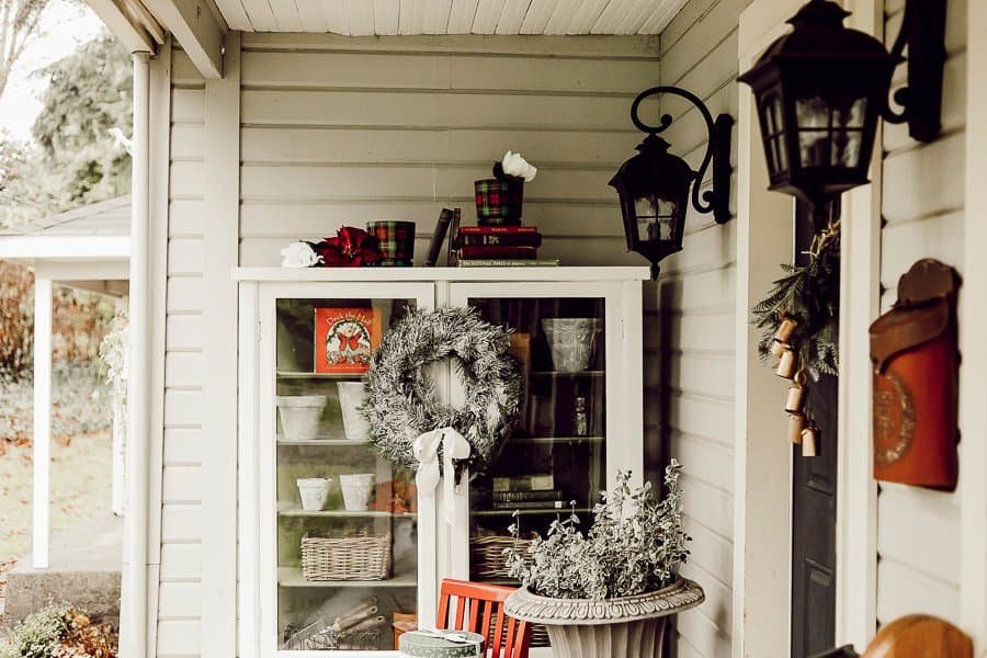 Red and green farmhouse Christmas porch with lanterns, a wreath and a painted garden cabinet