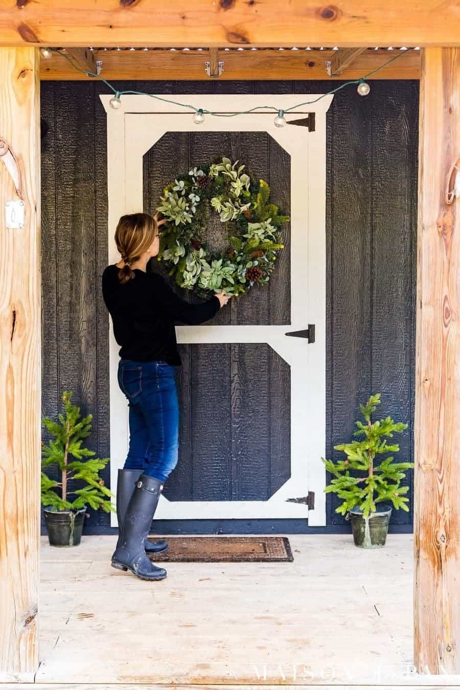 Black tiny house porch with a green wreath, small potted pine trees and globe string lights